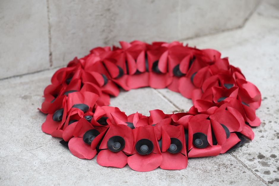 A floral wreath made of vibrant red petals and black centers, arranged in a circular shape, placed on a light-colored stone surface against a concrete wall. The wreath features fresh, softly textured 