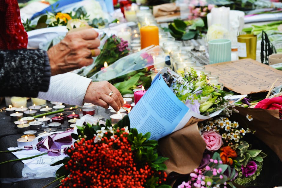A floral tribute scene showing hands arranging and holding bouquets of fresh flowers including roses, daisies, and greenery. The flowers are diverse in color, featuring pink, yellow, white, and purple