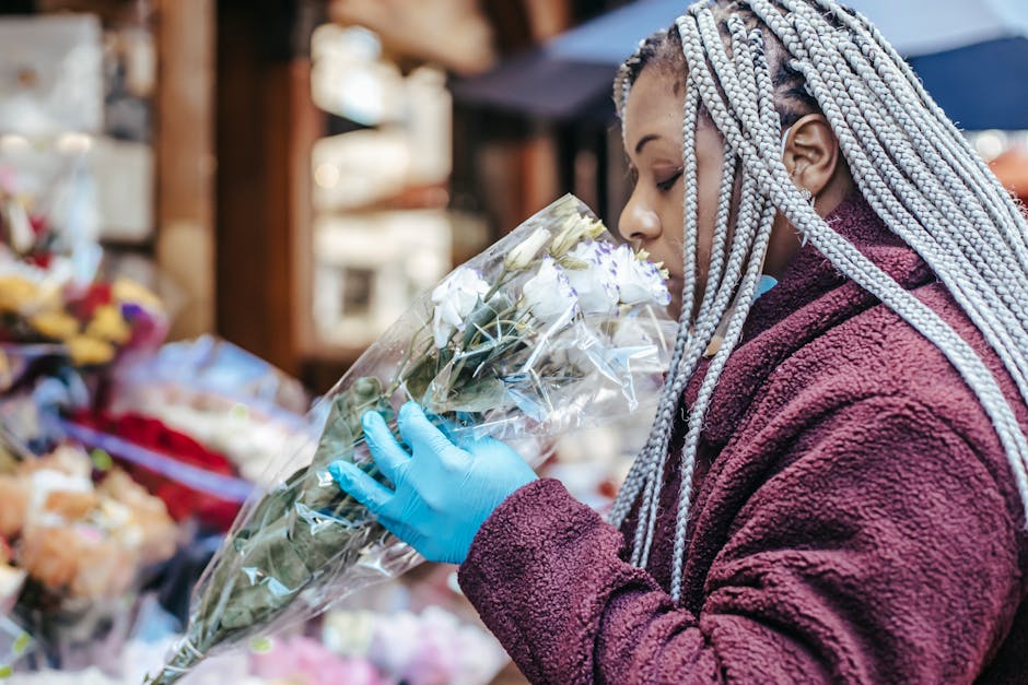 A woman with long, braided silver hair wearing a maroon fleece jacket and blue disposable gloves is holding a bouquet of white roses wrapped in transparent plastic. She is positioned in a flower shop,