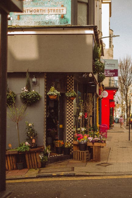 A floral shop facade displaying an array of fresh flowers in various arrangements outside the store, with potted plants and hanging baskets on the wall. The flowers include vibrant pink roses, white a
