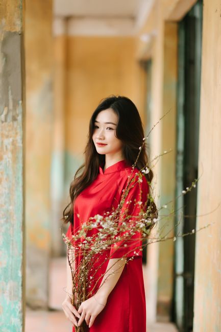 A young woman with long, dark hair and fair skin stands indoors, wearing a vibrant red silk dress with a high collar and short sleeves. She is holding a bouquet of mixed dried and fresh flowers, inclu